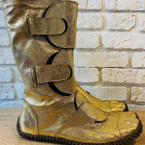 May include: Gold leather boots with a unique design featuring multiple straps and buckles. The boots have a rounded toe and a brown rubber sole.