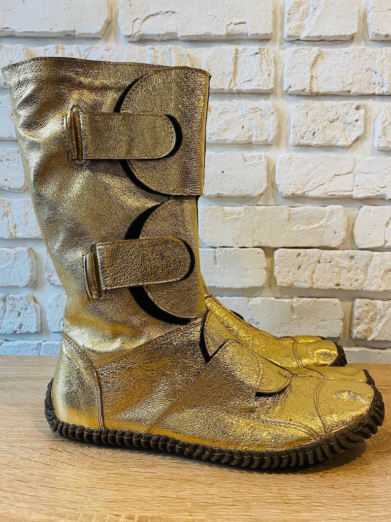 May include: Gold leather boots with a unique design featuring multiple straps and buckles. The boots have a rounded toe and a brown rubber sole.
