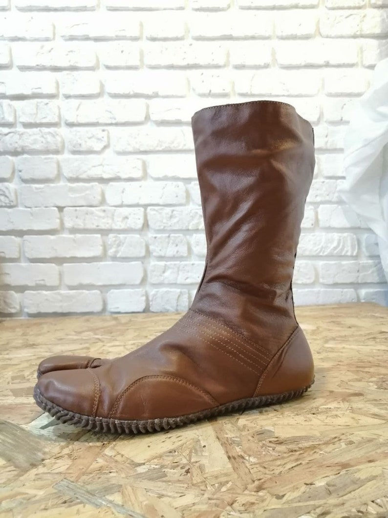 Puede incluir: Botas de cuero marr&oacute;n con un dise&ntilde;o de punta dividida. Las botas tienen un eje alto y un estilo simple y minimalista.