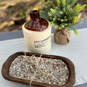 Può includere: Una natura morta rustica con una brocca in gres vintage con la parte superiore marrone e il testo "CITY BOTTLING CO. DENVER". Una ciotola di legno piena di pezzi di vetro iridescente legati con spago e un piccolo albero sempreverde finto in un sacco di juta.