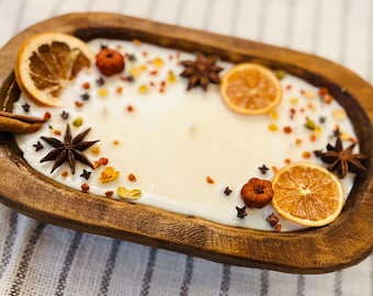 Könnte beinhalten: Eine große, cremefarbene Kerze in einer rustikalen Holzschale. Die Kerze ist mit getrockneten Orangenscheiben, Sternanis, Zimtstangen und kleinen dekorativen Kürbissen verziert. Die Schale ist etwa 30 cm lang.