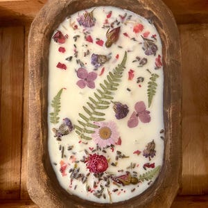 May include: A white candle with dried flowers and leaves embedded in the wax. The candle is in a wooden bowl with a rustic finish.