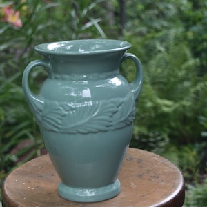 May include: A teal ceramic vase with two handles and a decorative leaf pattern. The vase has a wide opening and sits on a small pedestal base. The vase is approximately 10 inches tall.