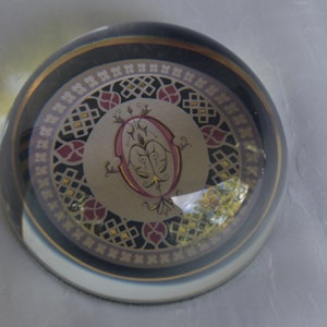 May include: A clear glass paperweight with a decorative design. The design features a central oval shape with a floral pattern in pink and gold, surrounded by a circular pattern in black, gold, and burgundy. The paperweight is dome-shaped.