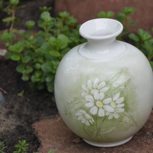 May include: A white ceramic vase with a round body and a narrow neck. The vase features a hand-painted design of white daisies with yellow centers and green leaves against a soft green background. The vase is displayed outdoors.