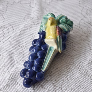 May include: A ceramic wall pocket shaped like a bunch of blue grapes with a yellow and red bird perched on top. The bird has green and white accents. Green leaves are behind the bird. The wall pocket is on a white lace tablecloth.