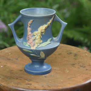 May include: A blue ceramic vase with two handles and a pedestal base. The vase features a floral design in yellow, pink, and green. The vase is sitting on a wooden stool.