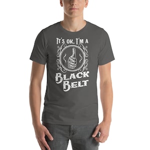 May include: A dark gray t-shirt with a white graphic design. The graphic features a thumbs up hand in a circle with the text "It's ok, I'm a Black Belt".