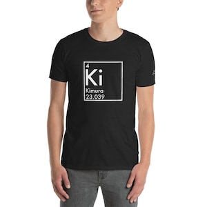 Pode incluir: Camiseta preta com um gráfico branco em estilo de tabela periódica que diz "Ki Kimura 23.039". A camiseta é de manga curta e tem gola redonda. A pessoa está usando calças cinzentas.