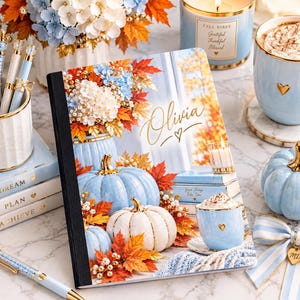 Puede incluir: Un cuaderno personalizado con una portada que muestra calabazas azul claro, hojas de otoño y una taza de chocolate caliente. El nombre Olivia está impreso en letras doradas. El cuaderno está rodeado de papelería a juego y una taza.
