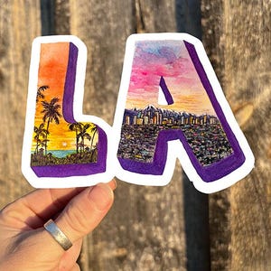 May include: A sticker with the letters "LA" in a 3D style. The "L" features a sunset scene with palm trees and the "A" features a cityscape with a sunset sky.