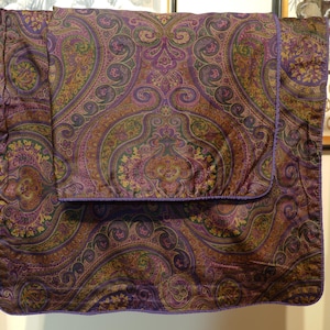 Könnte beinhalten: Ein Kissenbezug mit Paisley-Muster in Lila, Braun und Grün. Der Kissenbezug hat einen lila Rand und wird auf einem Kleiderständer ausgestellt. Das komplizierte Design zeigt wirbelnde Blumen- und Tropfenmotive.