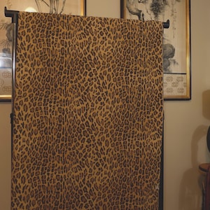 May include: A rectangular piece of fabric featuring a repeating leopard print pattern in shades of golden yellow, brown, and black. The textile is draped over a dark metal rod, displaying the full length of the animal-inspired design.