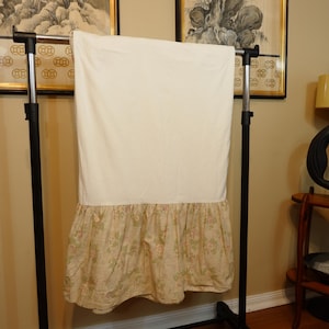 May include: A bed skirt featuring a solid cream-coloured top panel and a gathered bottom ruffle. The ruffle is decorated with a repeating floral pattern in muted shades of pink, green, and tan, creating a soft contrast against the plain fabric.