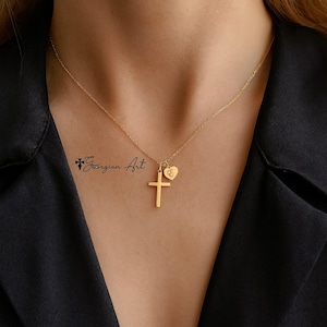May include: A gold necklace with a cross pendant and a heart charm. The necklace is worn against a black top. The chain is visible against the skin. The image also shows the text "Georgian Art".