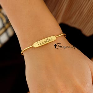 May include: A gold-colored bangle bracelet with the name "Nicolas" engraved on a rectangular plate. The bracelet is simple and elegant, with a delicate design. The image also includes the text "Georgian Art".