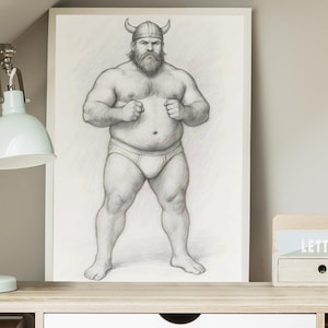 Puede incluir: Un dibujo a lápiz enmarcado de un hombre musculoso con un casco con cuernos y calzoncillos. La obra de arte se exhibe en un escritorio de madera clara, junto a una lámpara de escritorio verde menta y un pequeño sujetaletas de madera.