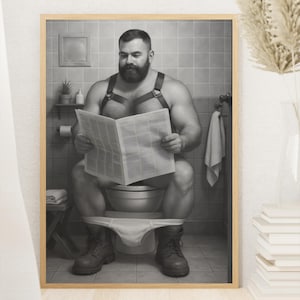 May include: Black and white framed print of a man with a beard reading a newspaper while sitting on a toilet. The man is wearing a leather harness and boots. The bathroom has a tiled wall and a small shelf with a plant.