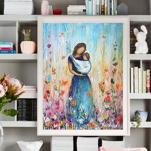 May include: A framed painting of a woman holding a child in a field of flowers. The painting is in a white frame and is hanging on a wall in a room with a pink chair and a bookshelf.