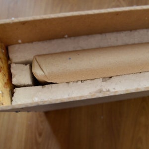 Puede incluir: Un tubo de cart&oacute;n envuelto en papel marr&oacute;n est&aacute; asegurado dentro de una caja de madera con acolchado de espuma blanca.