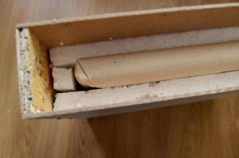 Puede incluir: Un tubo de cart&oacute;n envuelto en papel marr&oacute;n est&aacute; asegurado dentro de una caja de madera con acolchado de espuma blanca.