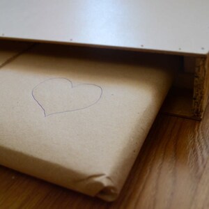 Puede incluir: Un paquete de papel marr&oacute;n con un coraz&oacute;n dibujado a mano en la parte superior. El paquete est&aacute; envuelto en cuerda y est&aacute; sobre una superficie de madera.