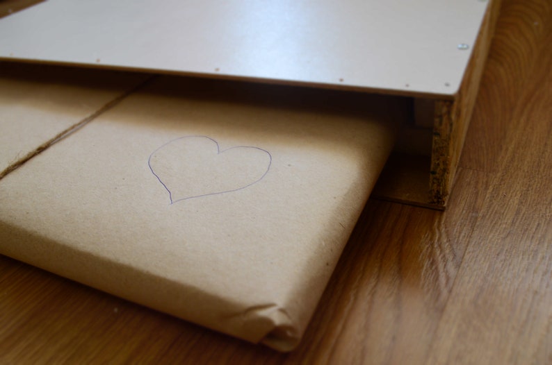 Pu&ograve; includere: Un pacco di carta marrone con un cuore disegnato a mano sopra. Il pacco &egrave; avvolto con spago e si trova all'interno di una scatola di legno.