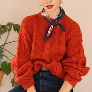 Puede incluir: Un suéter naranja tejido con trenzas, con cuello redondo y mangas largas. El suéter se combina con vaqueros azul oscuro y una bufanda estampada. Se ve un cinturón marrón con hebilla dorada. La textura y el color del suéter son prominentes.