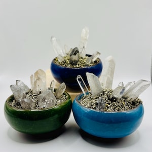 Könnte beinhalten: Drei kleine, glasierte Keramikschalen in Grün-, Blau- und Türkistönen. Jede Schale ist mit kleinen, dunklen Kieselsteinen und Clustern aus klaren Quarz Kristallen gefüllt. Die Schalen sind auf einer hellen Oberfläche angeordnet.