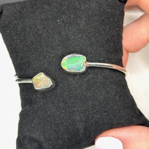 May include: A silver bangle bracelet with two opal stones. The stones are set in silver and are a light green and white color.
