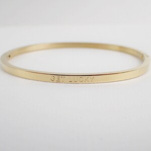 Puede incluir: Un brazalete de oro con las palabras "Get Lucky" grabadas en la parte delantera.