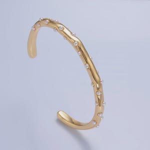 May include: A gold-colored open cuff bracelet with small, evenly spaced white pearls. The bracelet has a smooth, polished finish and a simple, elegant design. This jewelry piece is suitable for everyday wear or special occasions.