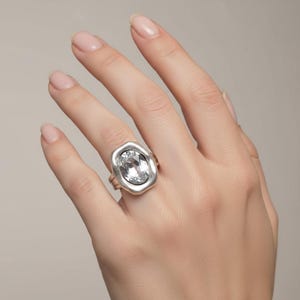 May include: A silver ring with a large, oval, clear gemstone. The ring has a thick, irregular bezel setting. The ring is on a hand with manicured nails.