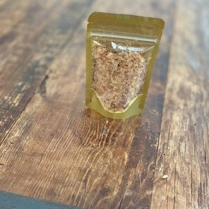 以下が含まれることがあります： 透明なプラスチック窓が付いた小さな金のポーチで、薄い茶色の細かく刻まれた食品が入っています。