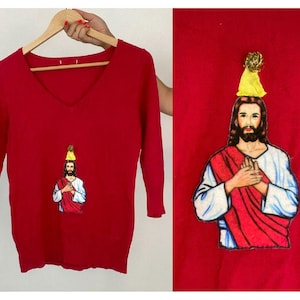 Könnte beinhalten: Roter Strickpullover mit V-Ausschnitt und einem Motiv von Jesus mit einer Partymütze.