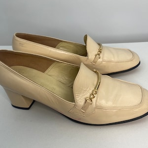 May include: A pair of cream-colored leather heeled loafers with a gold chain detail across the vamp.