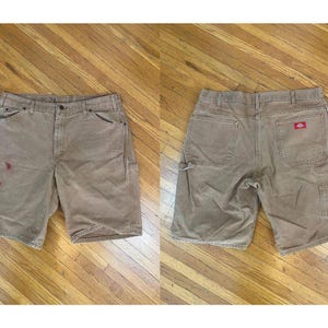 May include: A pair of brown work shorts with a red logo on the right back pocket. The shorts have two front pockets, two back pockets, and two side pockets.