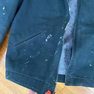 May include: A dark green work jacket with a red and white logo that says "Charter Construction". The jacket has a zipper and a pocket on the left side. The jacket is worn and has white paint splatters on it.