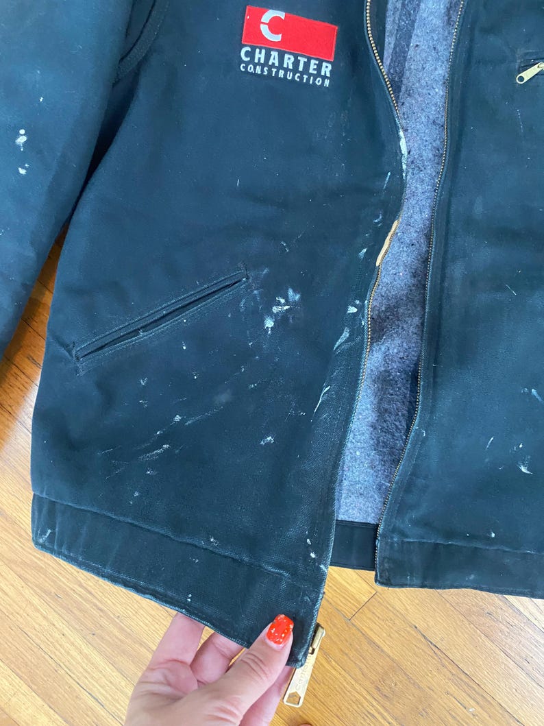 May include: A dark green work jacket with a red and white logo that says "Charter Construction". The jacket has a zipper and a pocket on the left side. The jacket is worn and has white paint splatters on it.