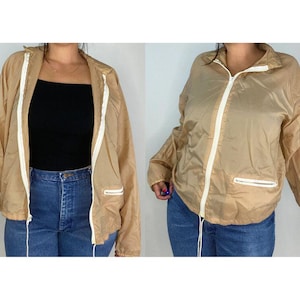 May include: A beige lightweight jacket with a zipper closure and white trim. The jacket has a zippered pocket on the left side.