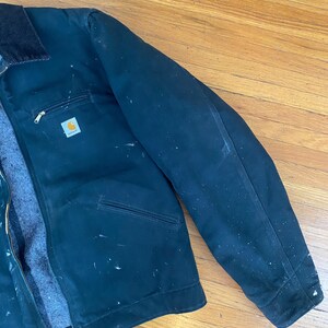 May include: A dark green work jacket with a zip fastening and a Carhartt logo patch on the left chest. The jacket has a corduroy collar and multiple pockets. The jacket is slightly worn and has some white paint splatters on it.