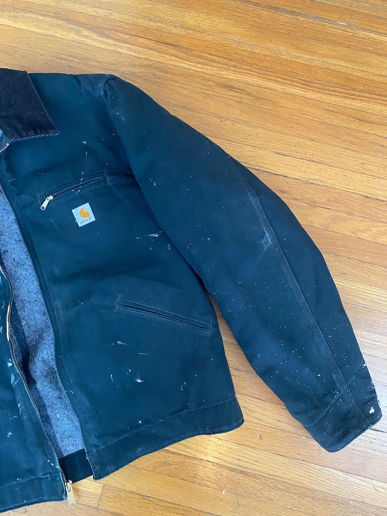 May include: A dark green work jacket with a zip fastening and a Carhartt logo patch on the left chest. The jacket has a corduroy collar and multiple pockets. The jacket is slightly worn and has some white paint splatters on it.