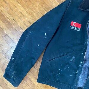 May include: A black work jacket with white paint splatters. The jacket has a red patch with a white letter "C" and the text "Charter Construction".