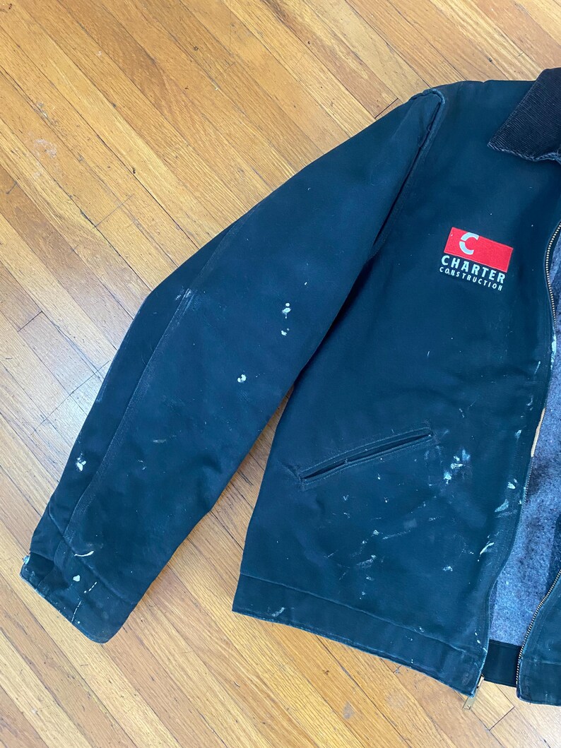 May include: A black work jacket with white paint splatters. The jacket has a red patch with a white letter "C" and the text "Charter Construction".