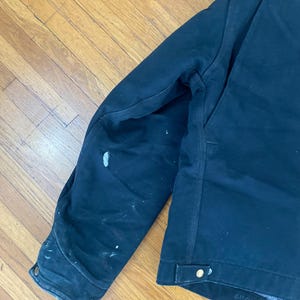May include: A dark blue work jacket with a zip fastening and a single button on the left side. The jacket has a worn look with white paint splatters on the left sleeve. The size tag on the jacket reads "61C39".