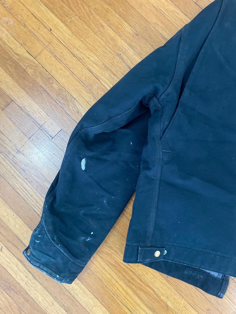 May include: A dark blue work jacket with a zip fastening and a single button on the left side. The jacket has a worn look with white paint splatters on the left sleeve. The size tag on the jacket reads "61C39".