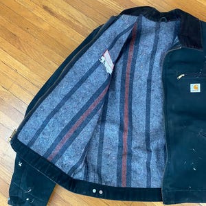 May include: A black Carhartt work jacket with a striped flannel lining. The jacket has a zip closure and a corduroy collar.