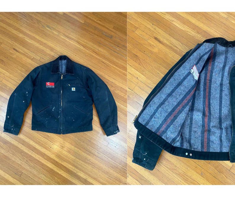 May include: A black work jacket with a Carhartt logo and a red patch on the left chest. The jacket has a zip fastening and two pockets on the front. The inside of the jacket is lined with a striped fabric.
