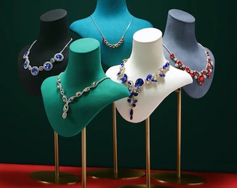 May include: Five velvet-covered bust forms on gold stands display a variety of necklaces with colorful gemstones. The bust forms are in shades of teal, black, green, white, and gray.