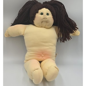 May include: A soft-sculpture doll with a pale fabric body, a prominent belly button, and dark brown yarn hair styled in two pigtails. The doll features embroidered green eyes, a small nose, and a stitched mouth, with a rounded head and limbs.